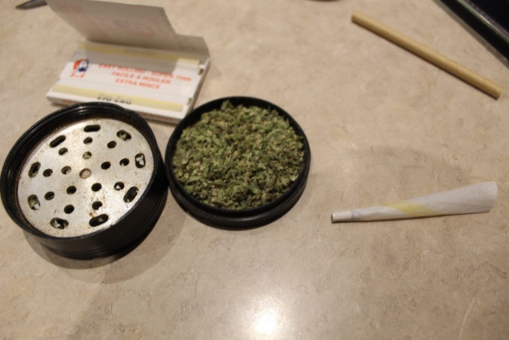 rolled joint beside a grinder and package of rolling papers. The grinder is filled completely with cannabis.