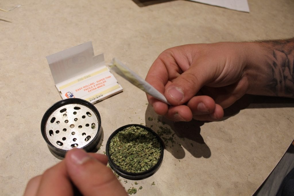 rolled joint being packed with more cannabis. Grinder full of weed and rolling papers on table beside them.