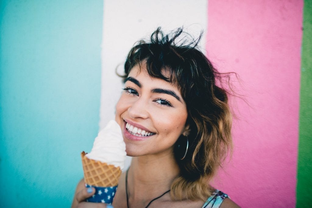 Woman holding a cannabis-infused ice cream cone smiling. Blue white and pink background. best cannabis edible recipes. 