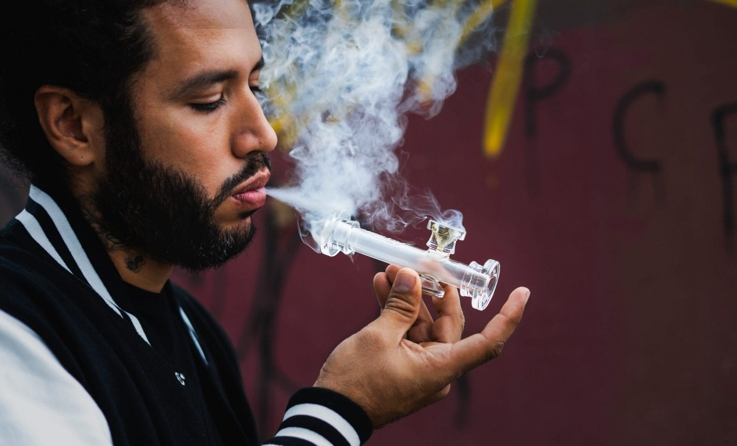 Man with beard smoking cannabis outside with a glass pipe. Smoke coming out of his mouth and the white pipe