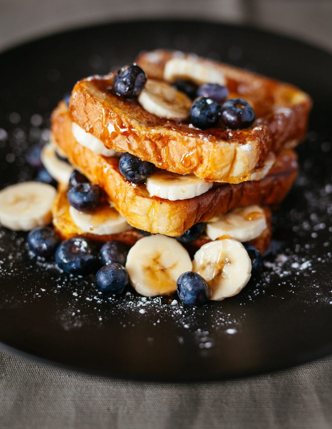 french toast on a black plate with blueberries, bananas, sugar