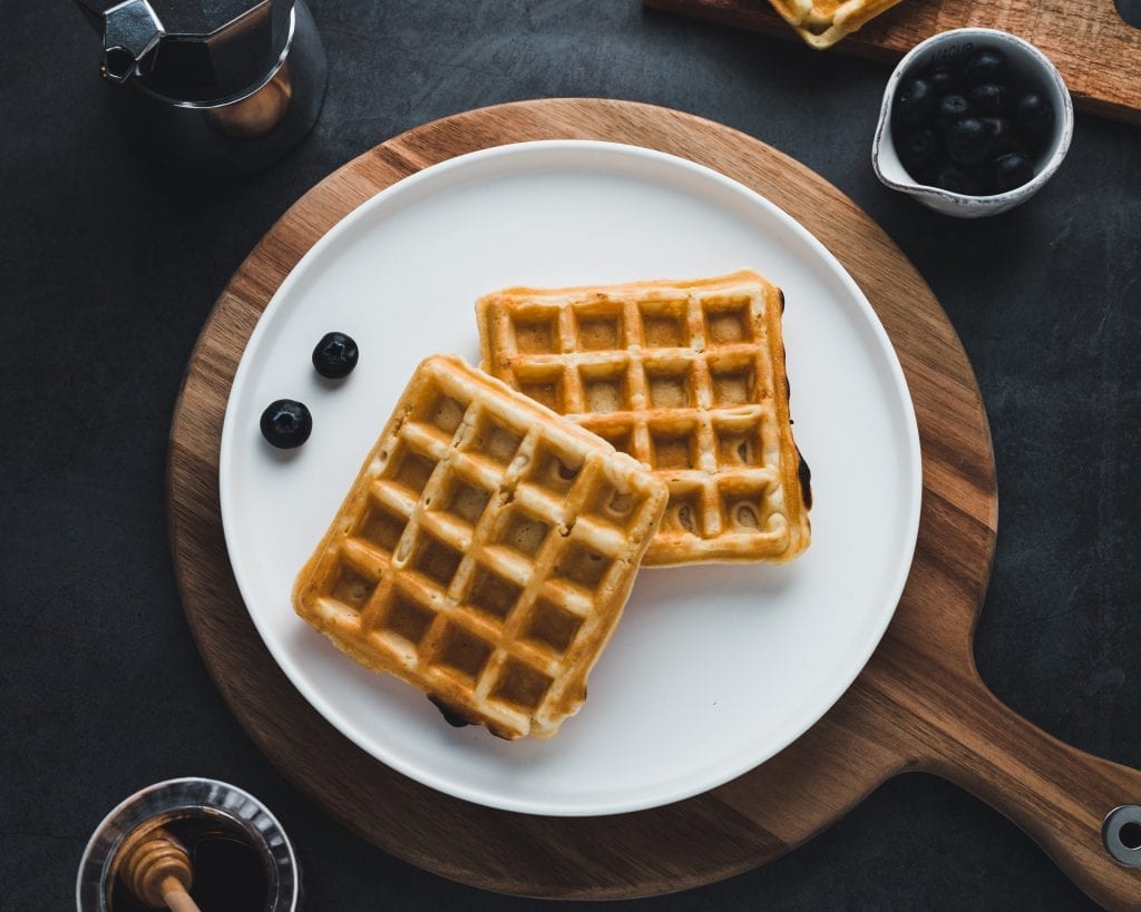 blueberry weed waffles on a white plate. the white plate has 2 blueberries on it. the plate is on a whie cutting board with honey and blueberries beside it on the table