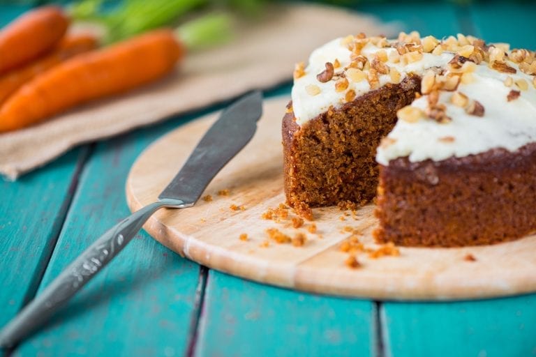 Weed carrot cake on a wooden cutting board with a slice taken out of it. Carrots in the background on a different cutting board