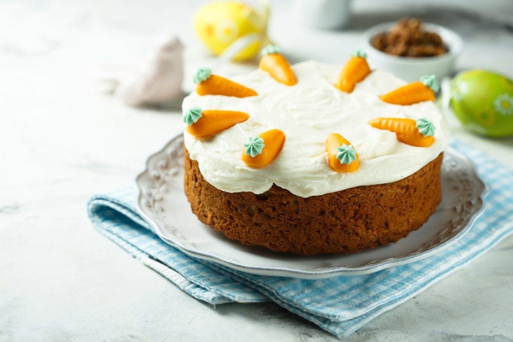 Cannabis infused carrot cake with white icing and decorative carrots on top. Other food in the background all on a white table