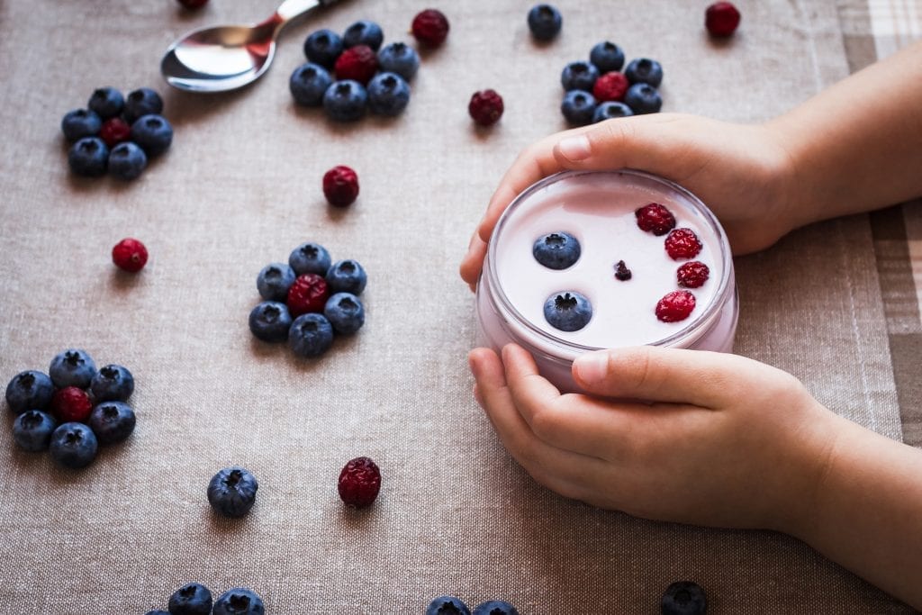 Child holding bowl of yogurt with blueberries and raspberries spread out on the table