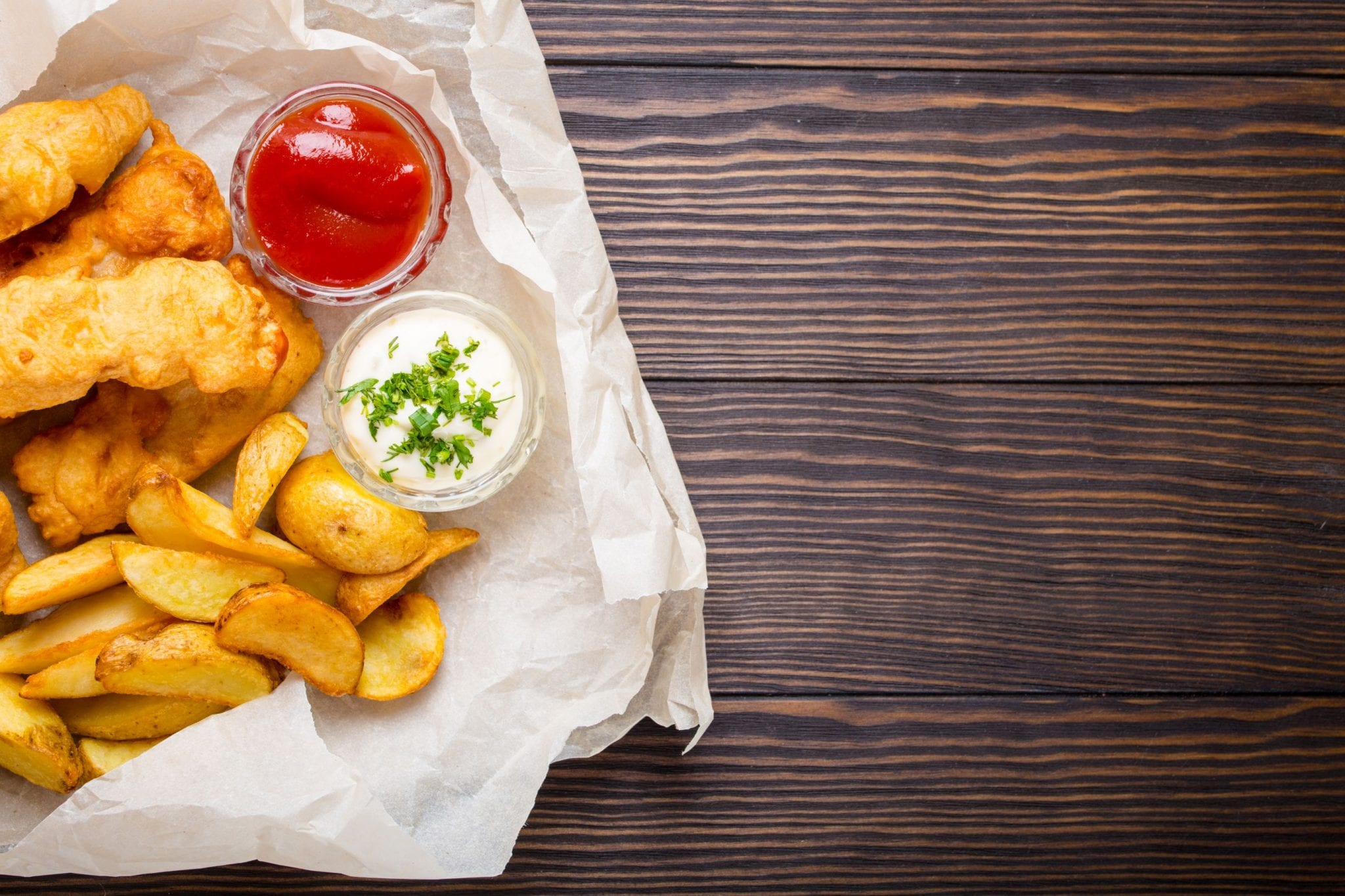 cannabis infused edible fish and chips on parchment paper with tarter sauce and ketchup beside them. All of this is on a wooden surface