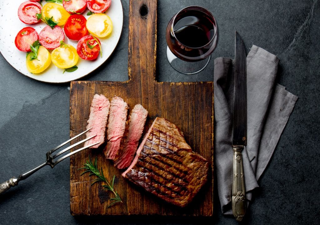 Weed steak with slices cut out of it on a cutting board with a knife and other cooking utensils beside it. There is a glass of wine and a plate of peppers in the background, A fancy weed edible dinner.