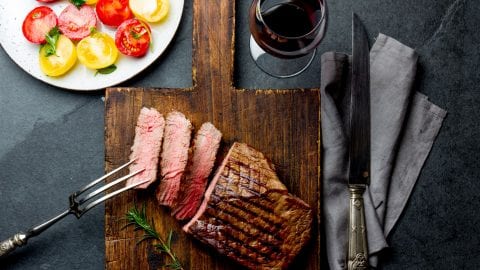 Weed steak with slices cut out of it on a cutting board with a knife and other cooking utensils beside it. There is a glass of wine and a plate of peppers in the background