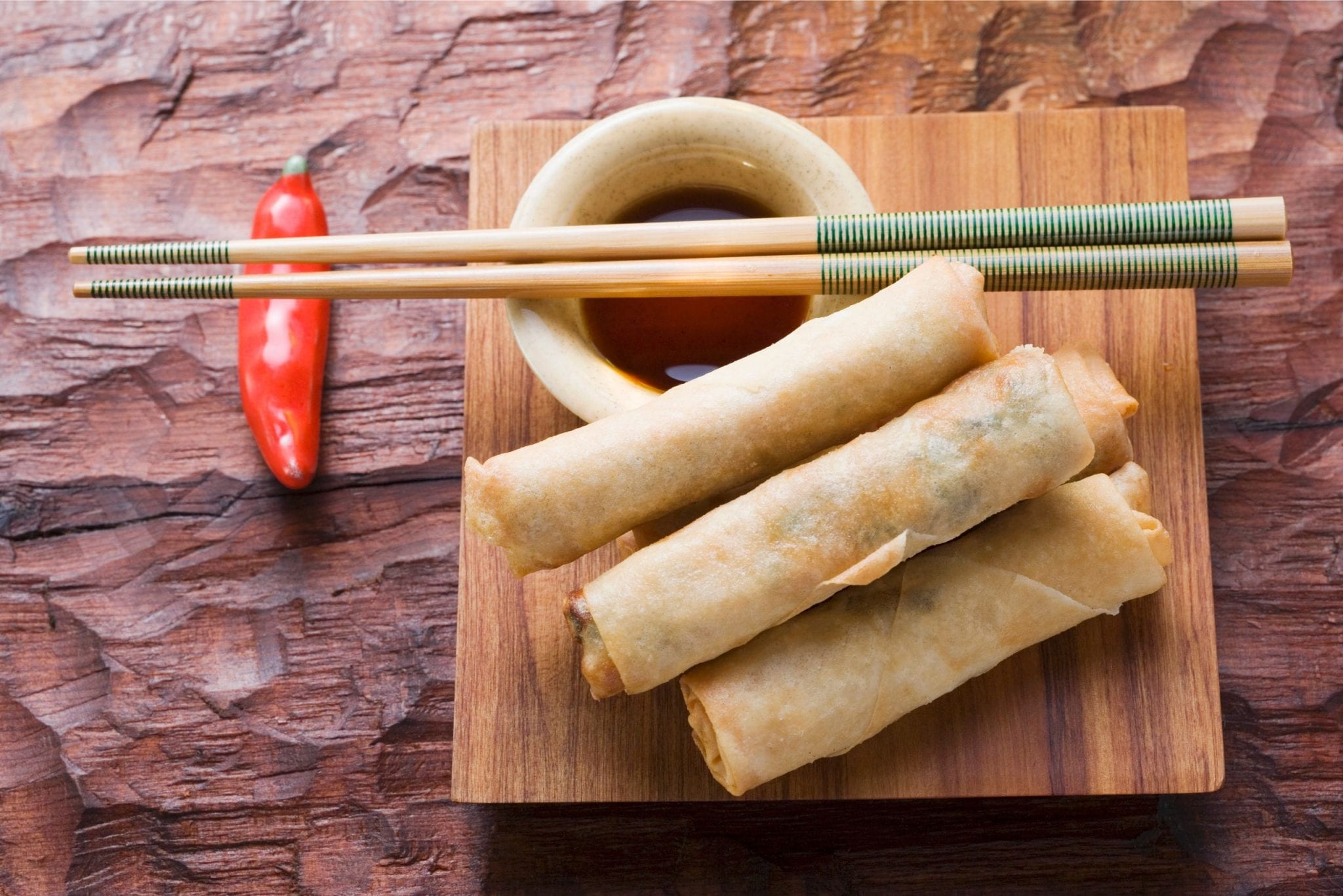 Three weed spring rolls on a wooden plate with a cup of sauce beside them. On top of the cup of sauce there is two chopsticks and a red pepper in the background