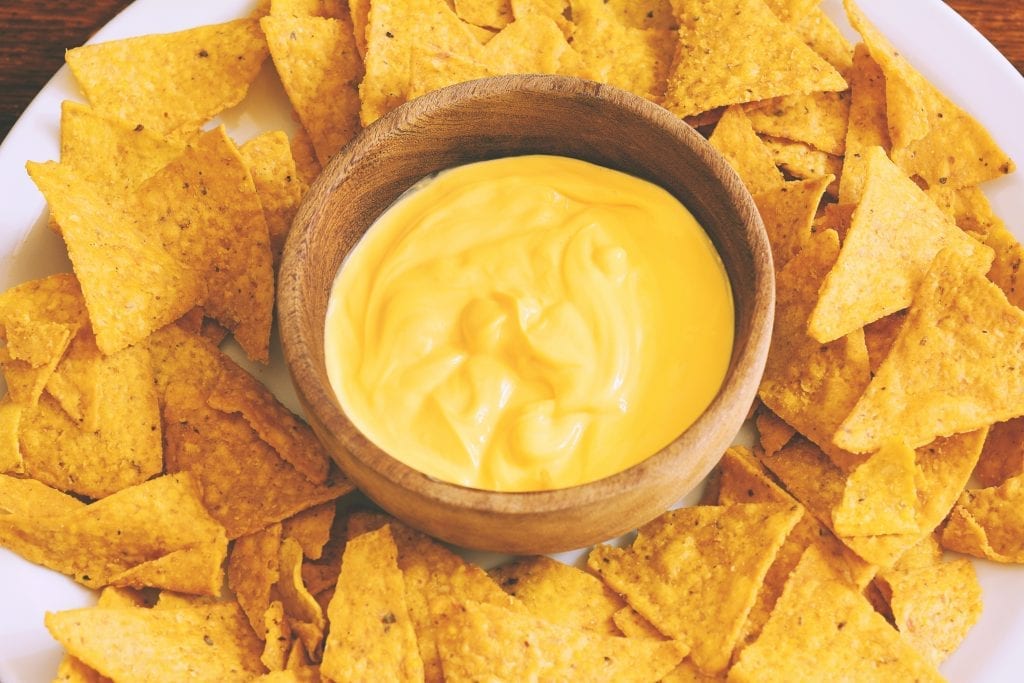 Top view of weed nacho cheese with tortilla chips spread around on a white plate. the plate is on a brown wooden table