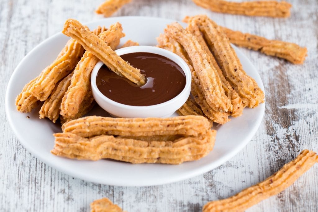 A white plate with lots of cannabis infused Churros on it with chocolate sauce in the middle of the plate that one churro is being dipped into