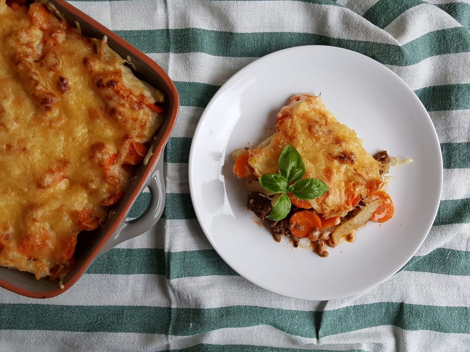 Breakfast casserole infused with cannabis in a cooking dish with a plate with a portion of the casserole on it all of this is on a striped kitchen towel