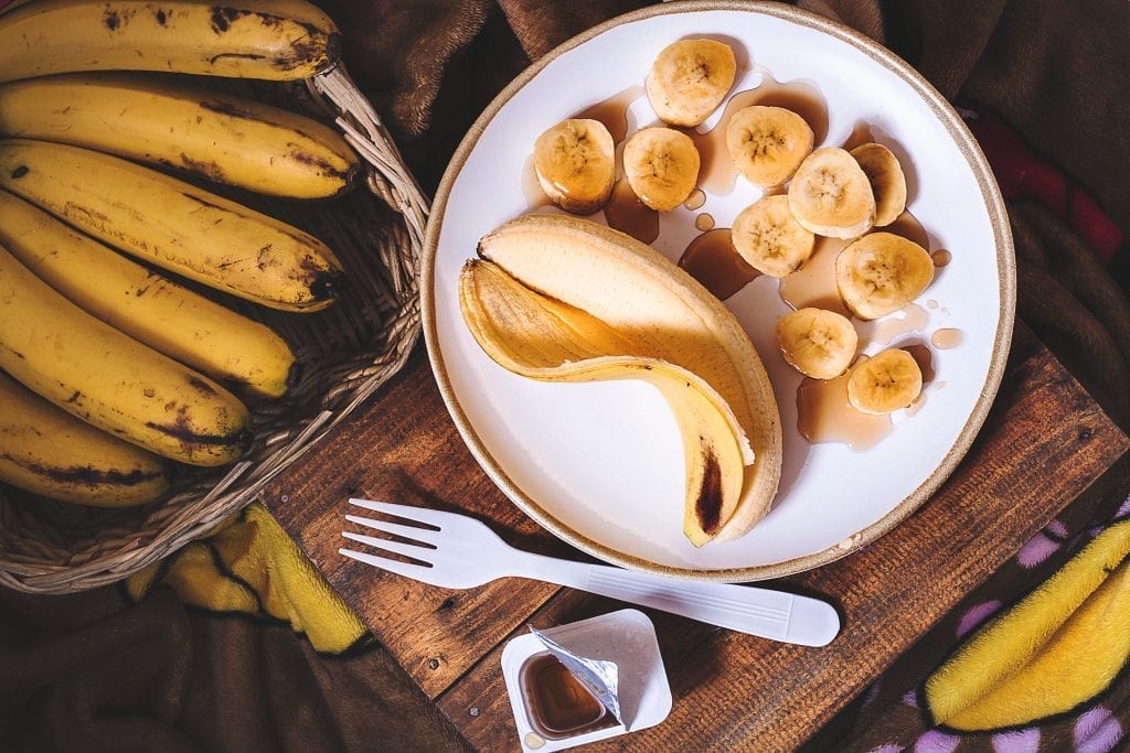 Banana on a plate that’s been cut open with another banana that still has the peel on it all of this is on a wooden cutting board with other bananas in the background