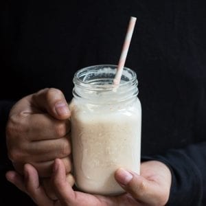 Edible banana milkshake with a pink straw in a person’s hands who is wearing a black sweater