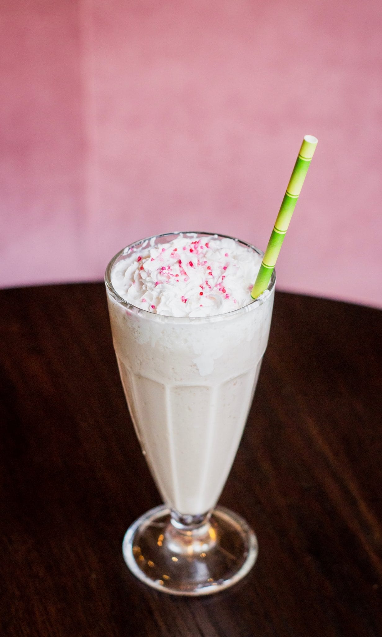 Weed milkshake with a green bamboo straw sticking out of it the milkshake has pink sprinkles on top of it and all of this is on a wooden table