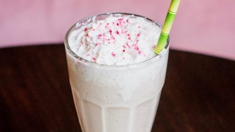 Weed milkshake with a green bamboo straw sticking out of it the milkshake has pink sprinkles on top of it and all of this is on a wooden table