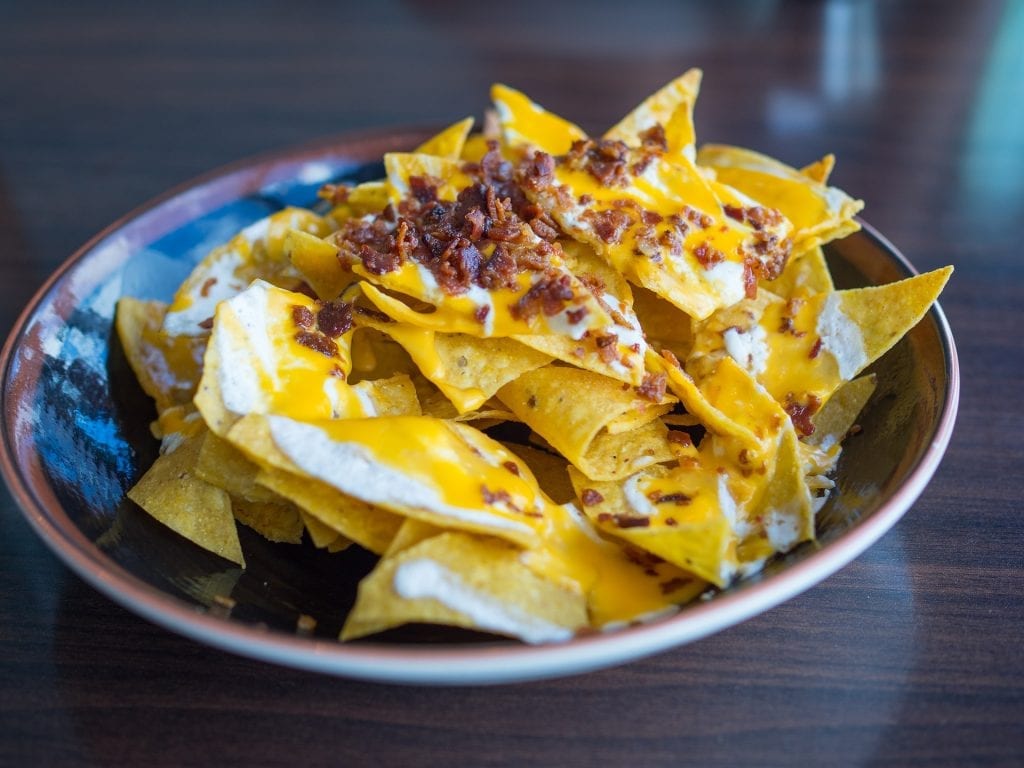 Bowl on a wooden table with nachos in it cannabis infused nacho cheese spread out over the nachos with bacon bits on top of it.