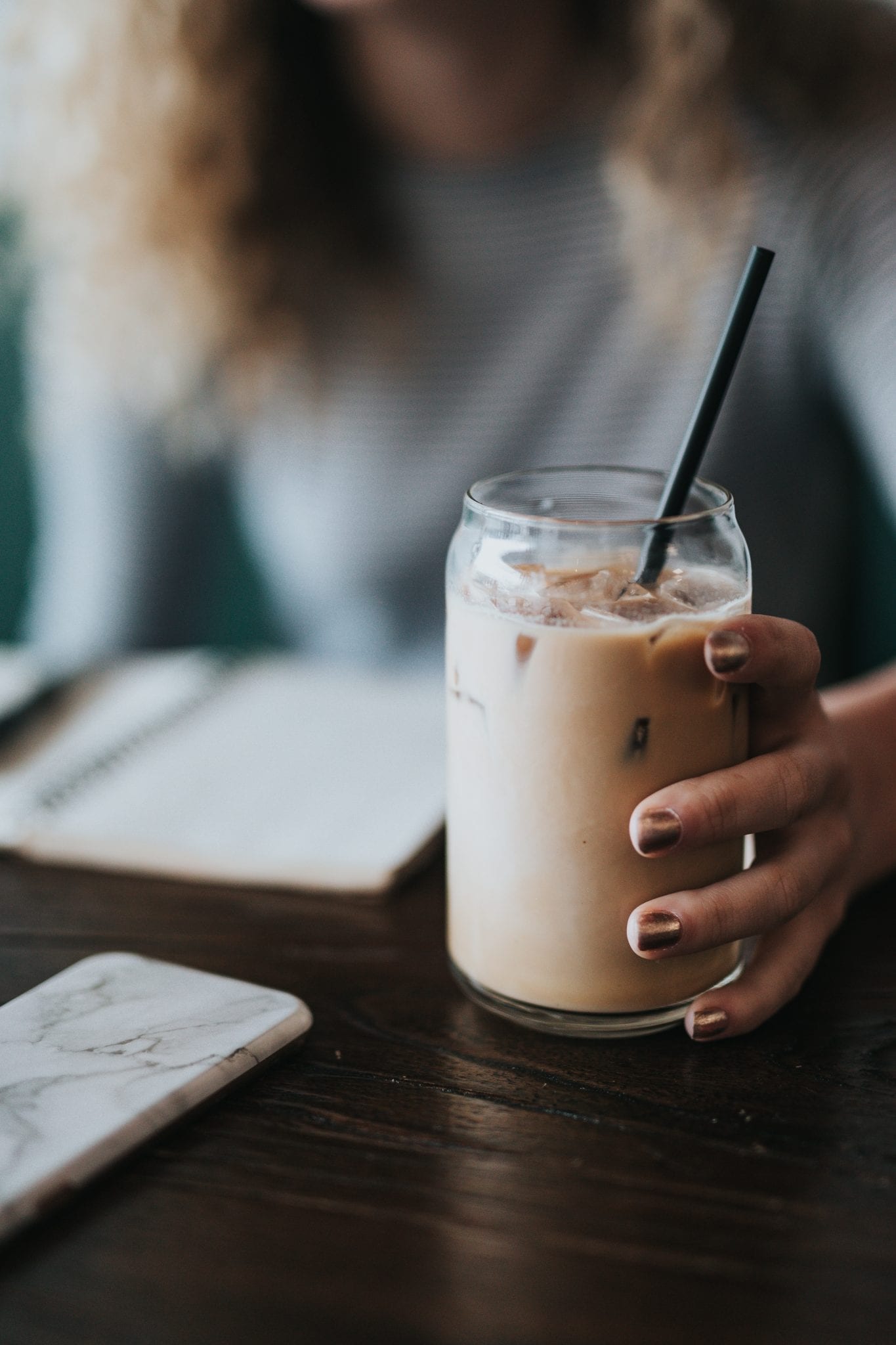 Woman holding jar with infused iced coffee in it with a black straw on a brown wooden table with notepad and iphone beside it