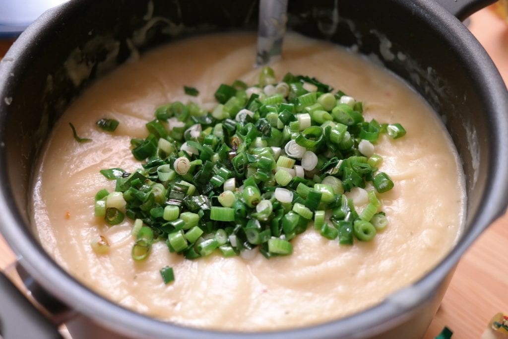 Cannabis potato soup in a pot with green onion on top of it the pot has a big spoon in it and its on a wooden cutting board