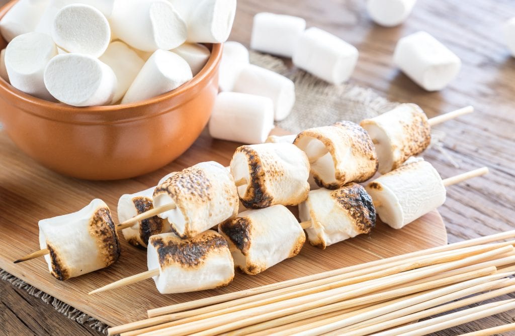 Cannabis infused Marshmallow skewers on the wooden board which is on a wooden surface with a bowl of weed marshmallows in the background