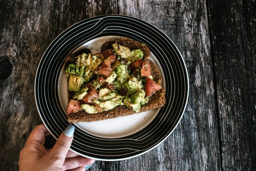 Weed infused avocado toast with tomatoes on a dinner plate. A ladies hand is holding the plate over a wooden table.