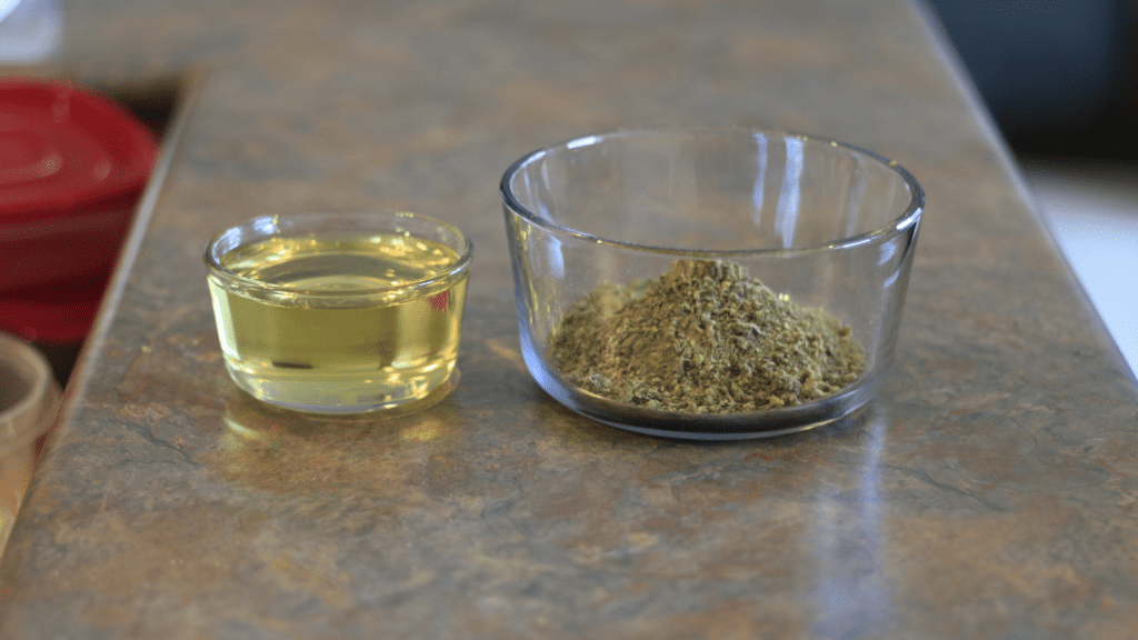 CBD-infused cooking oil that has a yellow colour to it, sits in a glass bowl on a counter top. A bow of ground weed sits beside it. 