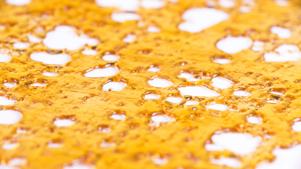 butane hash oil that is an amber colour sits on a white surface. The BHO resembles a honey or syrup texture.