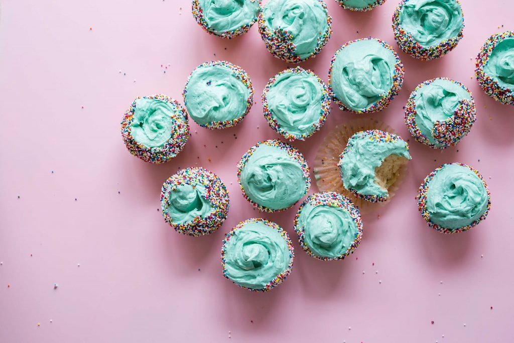 Gluten free weed cupcakes with light blue icing and rainbow sprinkles. The 16 cupcakes sit on a pink surface. 