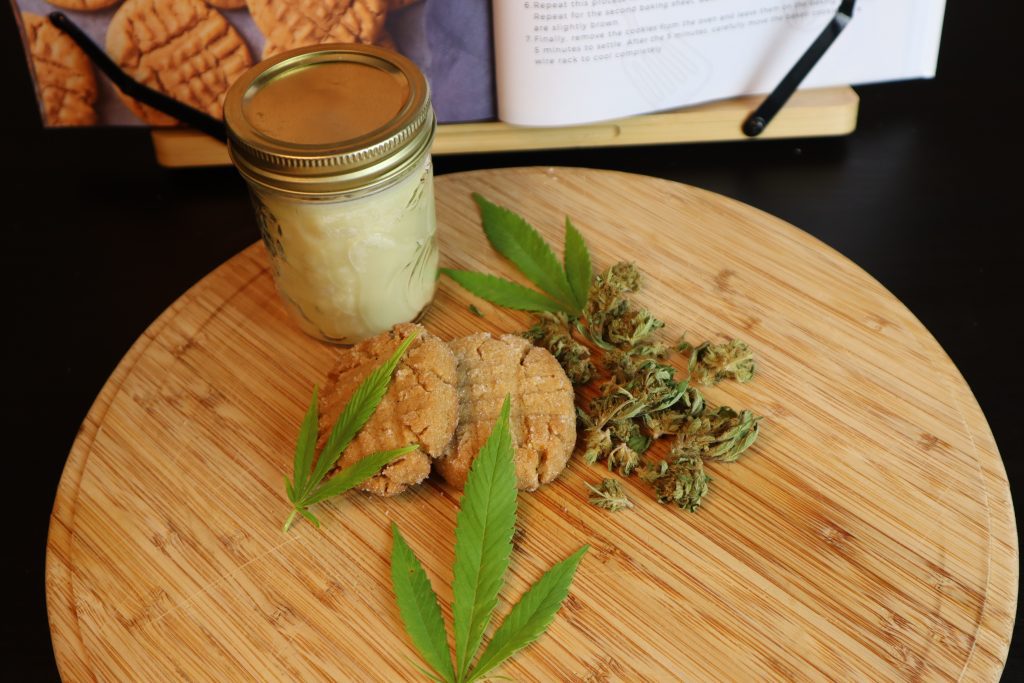 vegan weed peanut butter cookies on a cutting board with cannabis coconut oil, cannabis buds, and cannabis leafs beside it. A cannabis cookbook is in the background.